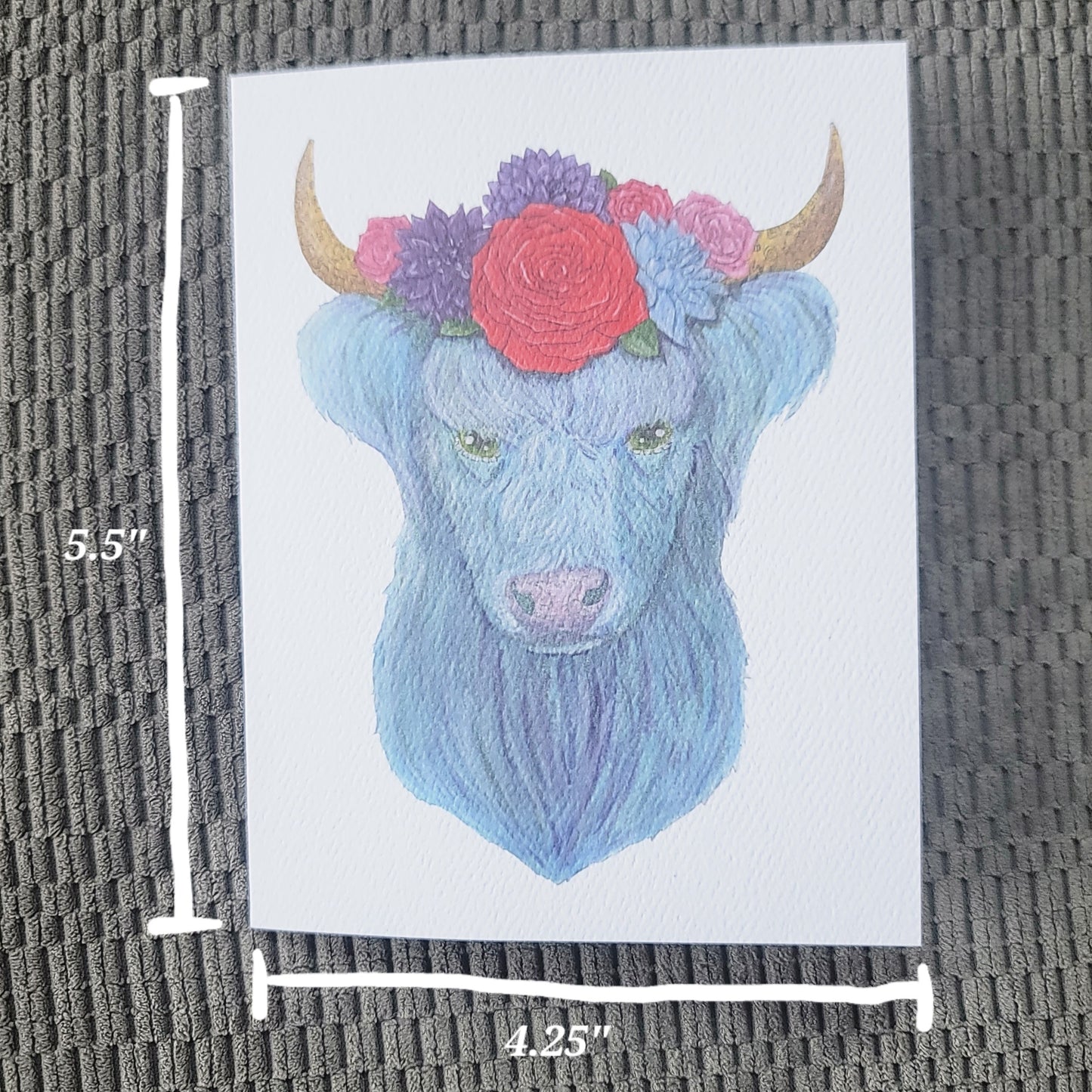 Watercolor illustration of a blue highland cow with a floral crown of daisies and roses on white card.