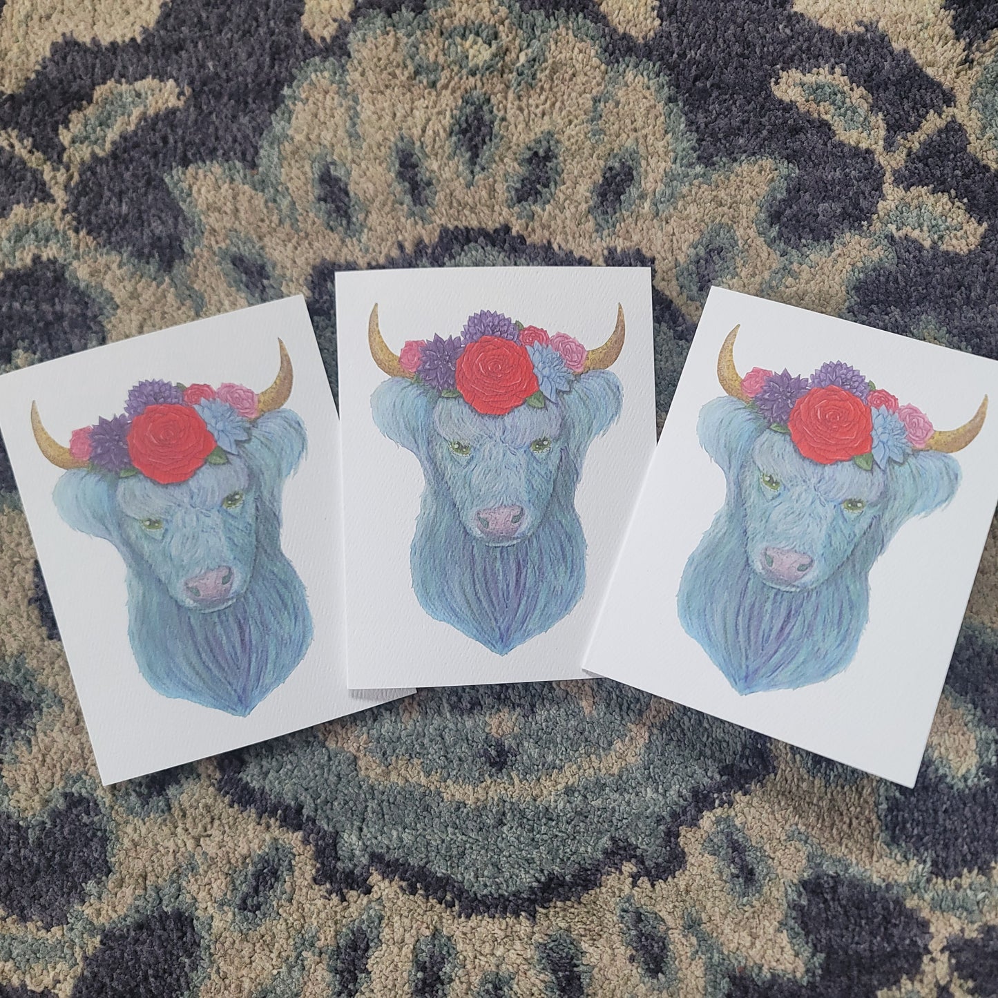 Three watercolor cards featuring a blue highland cow with flowers and horns on a patterned fabric background.