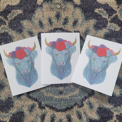 Three watercolor cards featuring a blue highland cow with flowers and horns on a patterned fabric background.