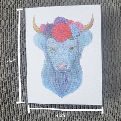 Watercolor illustration of a blue highland cow with a floral crown of daisies and roses on white card.